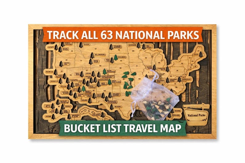 May include: Wooden map of the United States with the text "TRACK ALL 63 NATIONAL PARKS" and "BUCKET LIST TRAVEL MAP". The map is framed in a light wood color, with a dark wood background. Small green tree-shaped pins are included.