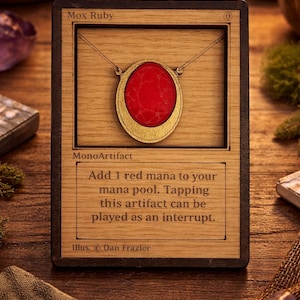 May include: Wooden Mox Ruby necklace pendant with a red stone set in a gold-colored frame. The pendant is displayed on a wooden card with text that reads "Mox Ruby" and "MonoArtifact".