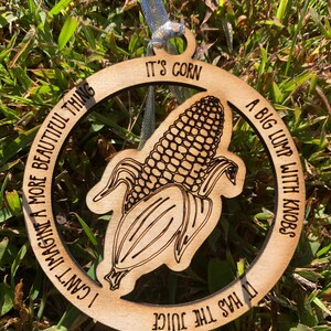 Puede incluir: Adorno de madera con un gr&aacute;fico de ma&iacute;z y el texto "IT'S CORN" y "A BIG LUMP WITH KNOBS". El adorno tambi&eacute;n incluye las frases "I CAN'T IMAGINE A MORE BEAUTIFUL THING" y "IT HAS THE JUICE". Tiene una cinta plateada para colgar.