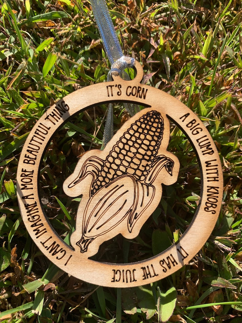 Puede incluir: Adorno de madera con un gr&aacute;fico de ma&iacute;z y el texto "IT'S CORN" y "A BIG LUMP WITH KNOBS". El adorno tambi&eacute;n incluye las frases "I CAN'T IMAGINE A MORE BEAUTIFUL THING" y "IT HAS THE JUICE". Tiene una cinta plateada para colgar.