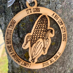 Puede incluir: Adorno de madera con un gr&aacute;fico de ma&iacute;z y el texto "It's Corn". El adorno tiene una cinta plateada para colgar. El texto alrededor del ma&iacute;z dice "I can't imagine a more beautiful thing" y "A big lump with knobs, it has the juice."