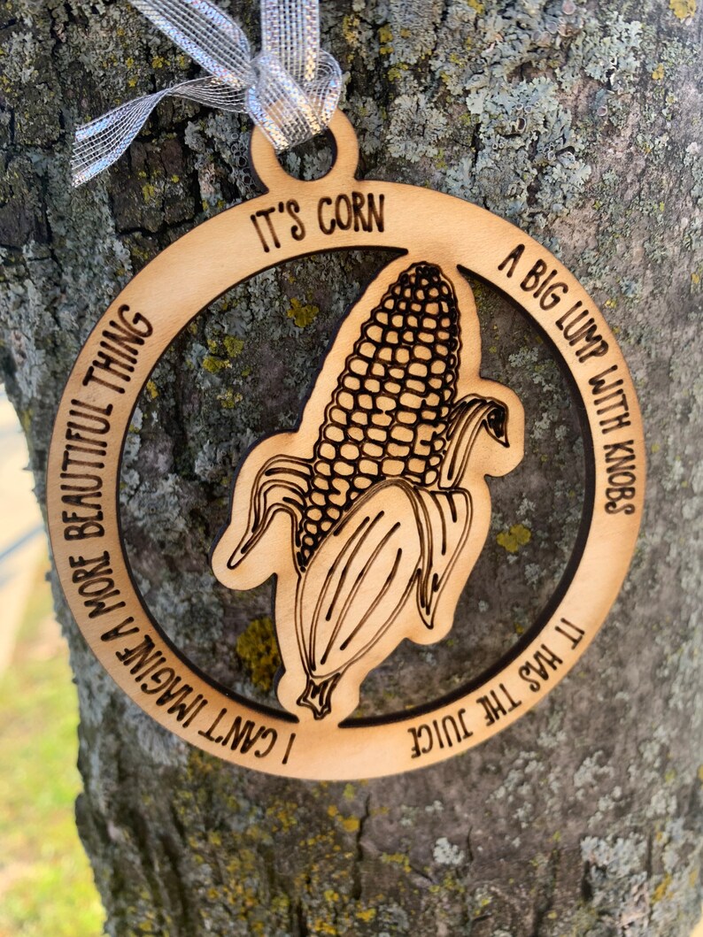 Puede incluir: Adorno de madera con un gr&aacute;fico de ma&iacute;z y el texto "It's Corn". El adorno tiene una cinta plateada para colgar. El texto alrededor del ma&iacute;z dice "I can't imagine a more beautiful thing" y "A big lump with knobs, it has the juice."