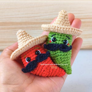 Pode incluir: Duas pimentas de crochê, uma vermelha e uma verde, cada uma usando um pequeno sombrero bege e um bigode preto. Ambas as pimentas possuem olhos de segurança de plástico preto e detalhes bordados. O texto AmigurumisMonterrey Etsy é visível.
