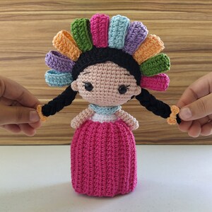 May include: A crocheted doll with black hair, a pink dress, and a colorful flower crown. The doll has large eyes and a sweet expression.