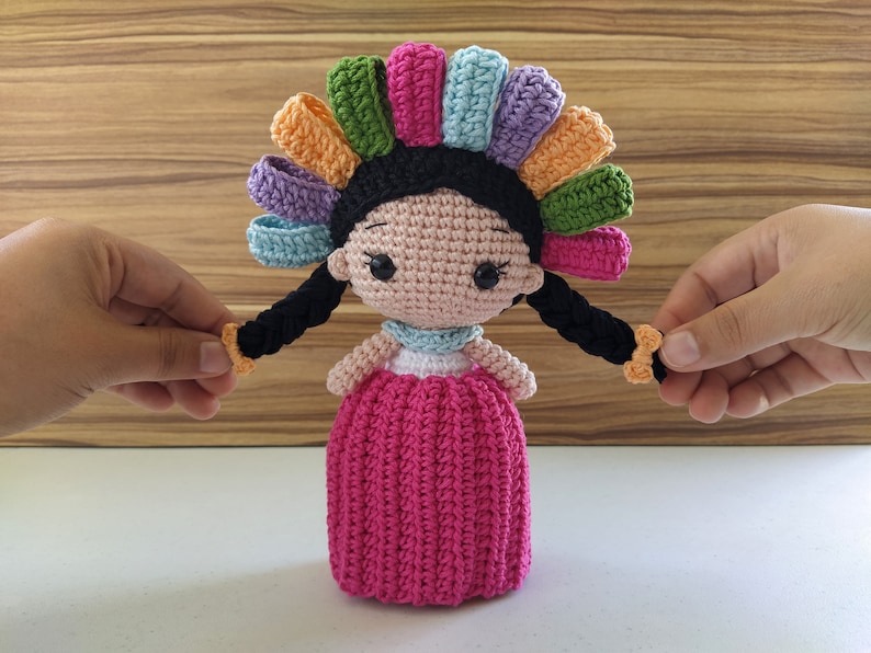 May include: A crocheted doll with black hair, a pink dress, and a colorful flower crown. The doll has large eyes and a sweet expression.