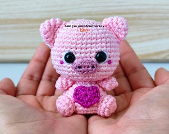 Könnte beinhalten: Ein handgehäkeltes rosa Ferkel mit schwarzen Sicherheitsaugen und einem lila Herz. Das Ferkel hat kleine Ohren und eine Schnauze. Der Text "AmigurumisMonterrey Etsy" ist im Hintergrund sichtbar.