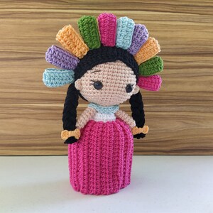 May include: A crocheted doll with black hair, a pink dress, and a colorful headpiece. The doll is handmade and has a sweet, whimsical expression.