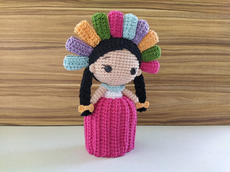 May include: A crocheted doll with black hair, a pink dress, and a colorful headpiece. The doll is handmade and has a sweet, whimsical expression.