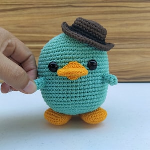 May include: A teal blue crocheted plush toy shaped like a duck wearing a brown felt cowboy hat. The duck has orange feet and a yellow beak.
