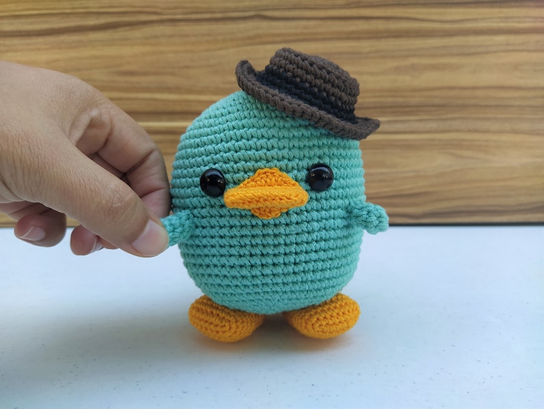 May include: A teal blue crocheted plush toy shaped like a duck wearing a brown felt cowboy hat. The duck has orange feet and a yellow beak.
