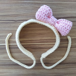 May include: A crocheted headband with a pink bow. The headband is cream colored and the bow is a light pink.