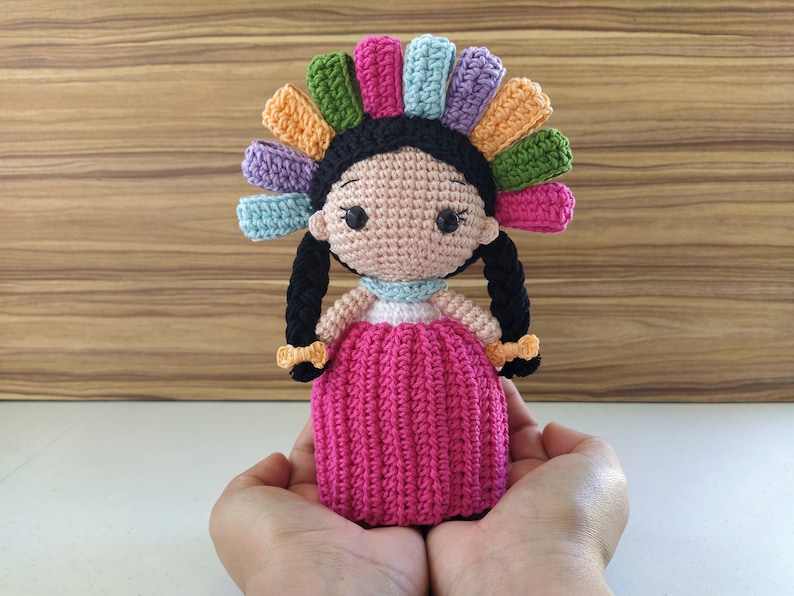 May include: A crocheted doll with black hair, a pink dress, and a colorful flower crown. The doll is being held in the palms of two hands.