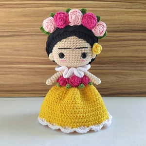May include: A crocheted doll with a yellow dress, white lace trim, and a flower crown. The doll has black hair, brown eyes, and a painted-on unibrow.