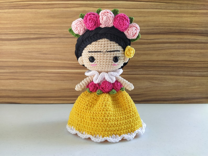 May include: A crocheted doll with a yellow dress, white lace trim, and a flower crown. The doll has black hair, brown eyes, and a painted-on unibrow.