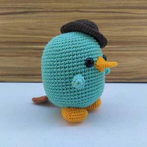 May include: A crocheted plush toy shaped like a platypus wearing a brown hat. The toy has a light blue body, orange beak and feet, and black eyes.