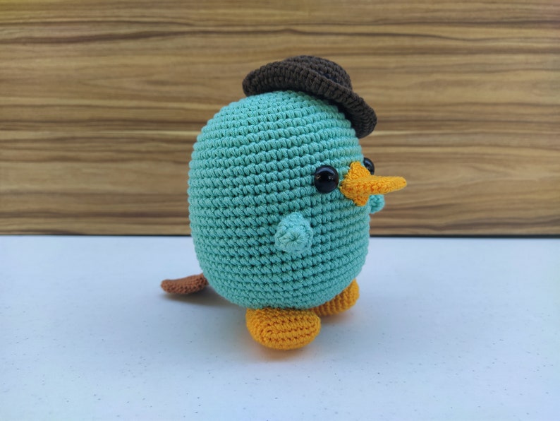 May include: A crocheted plush toy shaped like a platypus wearing a brown hat. The toy has a light blue body, orange beak and feet, and black eyes.