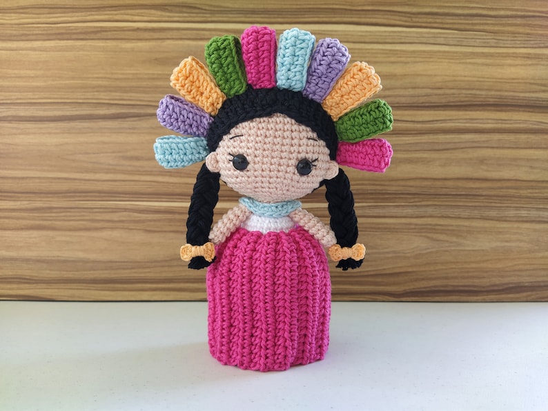 May include: A crocheted doll with a pink dress, black hair, and a colorful headpiece. The doll has a sweet expression and is handmade.