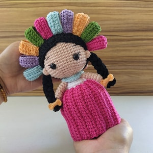 May include: A crocheted doll with a pink dress and a colorful headpiece. The doll has black hair in braids and brown eyes.