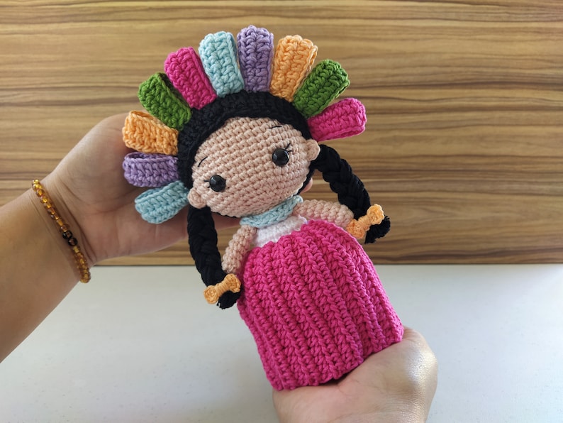 May include: A crocheted doll with a pink dress and a colorful headpiece. The doll has black hair in braids and brown eyes.