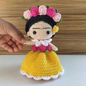 May include: Crocheted doll with black hair, a yellow dress, and a flower crown. The doll has a painted-on unibrow and mustache.