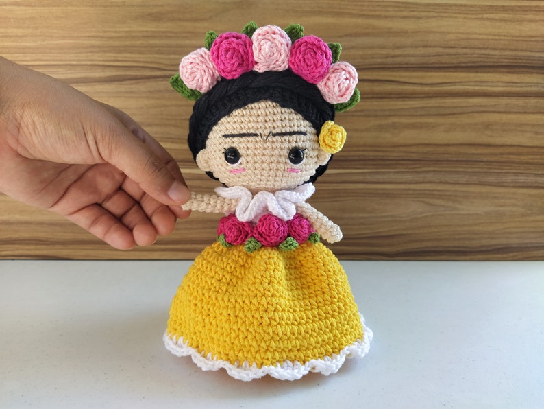 May include: Crocheted doll with black hair, a yellow dress, and a flower crown. The doll has a painted-on unibrow and mustache.