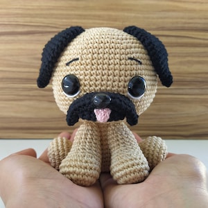 Puede incluir: Un animal de peluche de pug tejido a crochet con orejas negras, nariz negra y grandes ojos negros. El pug está sentado en las manos de una persona.