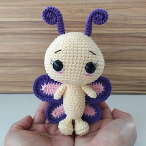 May include: A crocheted butterfly toy with purple wings and a yellow body. The butterfly has large black eyes and a pink nose.