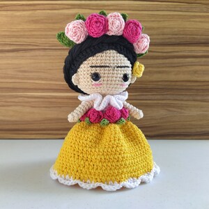 May include: A crocheted doll with black hair, a yellow dress, and a flower crown. The doll has a painted face with a unibrow and a small flower on her ear.