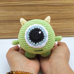 May include: A green crocheted monster toy with a large blue eye and white accents. The toy has two small horns on top of its head and is being held in the hands of a person.