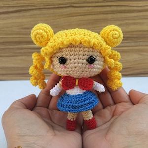 Könnte beinhalten: Eine gehäkelte Puppe mit gelben Haaren, einem weißen Shirt, einer roten Schleife, einem blauen Rock und roten Schuhen. Die Puppe hält einen weißen Zauberstab.