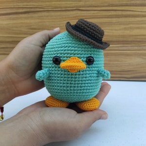 May include: A teal blue crocheted plush toy shaped like a duck wearing a brown felt hat. The toy has black eyes, an orange beak, and orange feet.