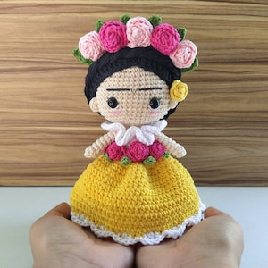 以下が含まれることがあります： 黒髪の毛糸のドールで、黄色のドレスを着ています。ピンクの花の冠をかぶっています。ドールは、少し眉をひそめた表情で、大きな目をしています。