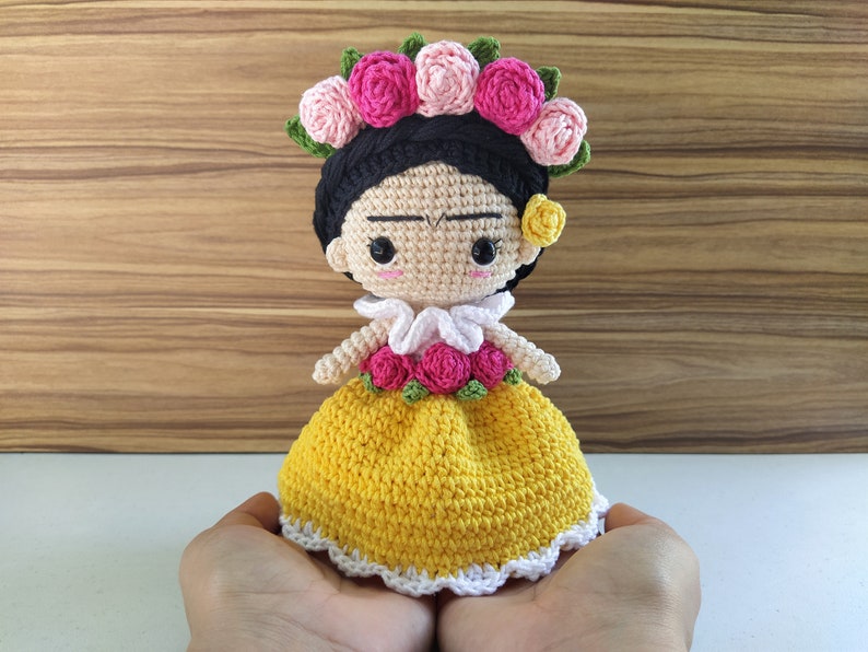 May include: A crocheted doll with black hair, a yellow dress, and a pink flower crown. The doll has a painted face with a slight frown and large eyes.