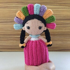 May include: A crocheted doll with black hair in braids, wearing a pink dress and a colorful headdress.