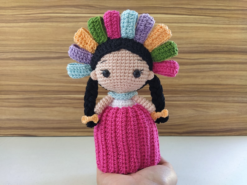 May include: A crocheted doll with black hair in braids, wearing a pink dress and a colorful headdress.