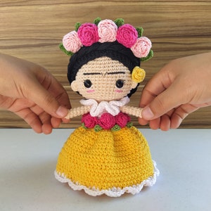 May include: A crocheted doll with a yellow dress, pink flowers, and a black braid. The doll is wearing a white collar and has a flower behind her ear.