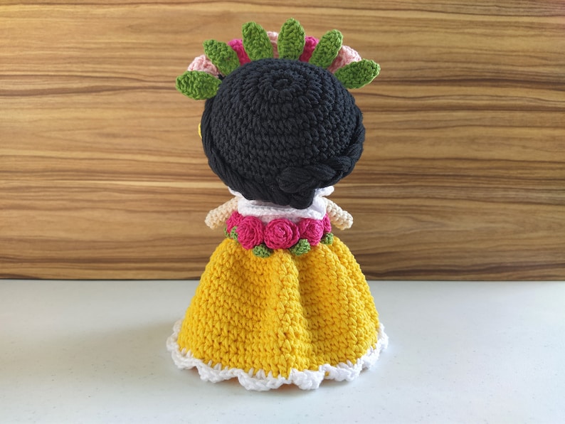 May include: A crocheted doll with black hair, a yellow dress with white trim, and a flower crown with pink and green flowers.