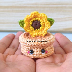 Puede incluir: Un girasol tejido a ganchillo en una maceta. La flor es amarilla con centro marrón y hojas verdes. La maceta es de color naranja claro con una cara sonriente y mejillas rosas. El logo de Etsy AmigurumisMonterrey es visible.