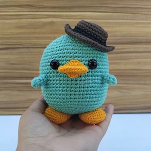 May include: A teal blue crocheted plush toy with a brown felt cowboy hat, orange beak, and black eyes. The toy has orange crocheted feet.