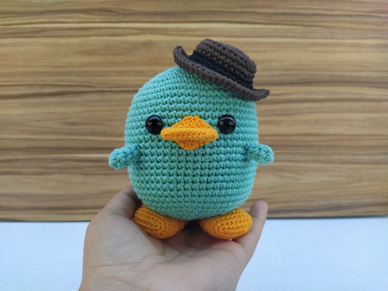May include: A teal blue crocheted plush toy with a brown felt cowboy hat, orange beak, and black eyes. The toy has orange crocheted feet.