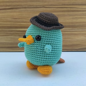 May include: A teal crocheted platypus wearing a brown felt hat with a wide brim. The platypus has orange feet and a yellow beak.