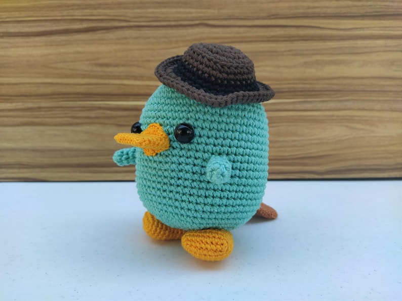 May include: A teal crocheted platypus wearing a brown felt hat with a wide brim. The platypus has orange feet and a yellow beak.