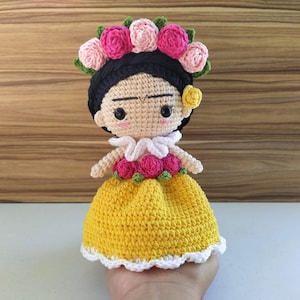 May include: Crocheted doll with black hair, a yellow dress, and a flower crown. The doll has a painted face with a unibrow and a small flower behind her ear.
