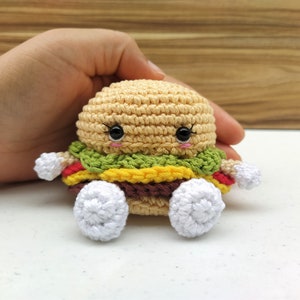 Può includere: Un giocattolo di peluche a forma di hamburger all'uncinetto con un viso sorridente, lattuga verde, formaggio giallo, pomodoro rosso e carne marrone. Il giocattolo ha piedi bianchi all'uncinetto.