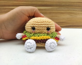 Op de afbeelding: Een gehaakt hamburger knuffel met een glimlachend gezicht, groene sla, gele kaas, rode tomaat en bruin vlees. Het speelgoed heeft witte gehaakte voeten.