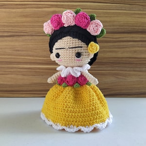 May include: A crocheted doll with black hair, a yellow dress, and a pink flower crown. The doll has a painted face with brown eyes and a slight frown.