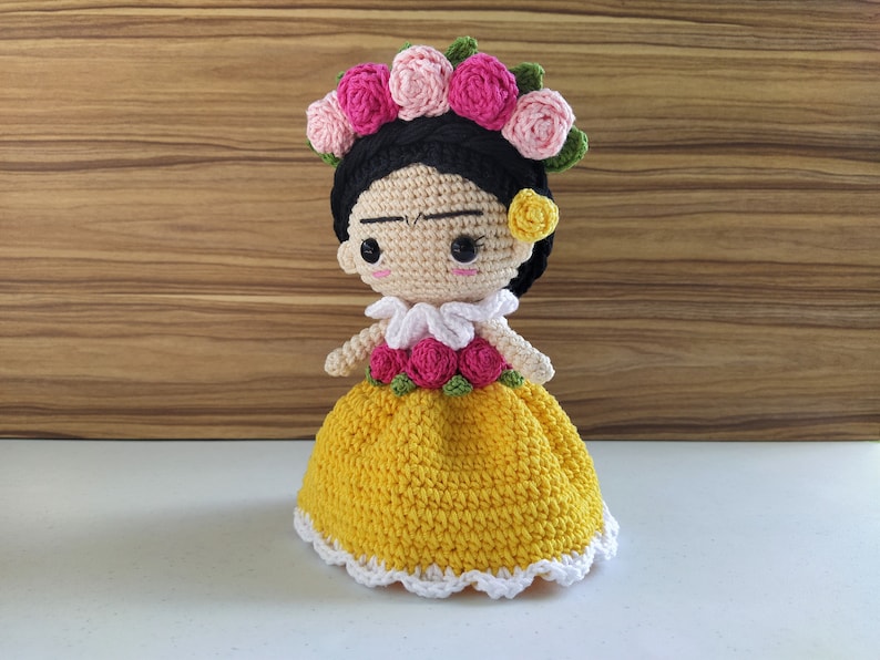 May include: A crocheted doll with black hair, a yellow dress, and a pink flower crown. The doll has a painted face with brown eyes and a slight frown.