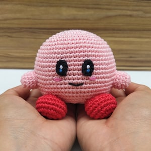 Peut inclure: Peluche en crochet rose avec des yeux noirs, une bouche souriante et des pieds en crochet rouge.
