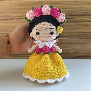 May include: A crocheted doll with black hair, a yellow dress, and a flower crown. The doll has a white collar and a pink flower on her ear.