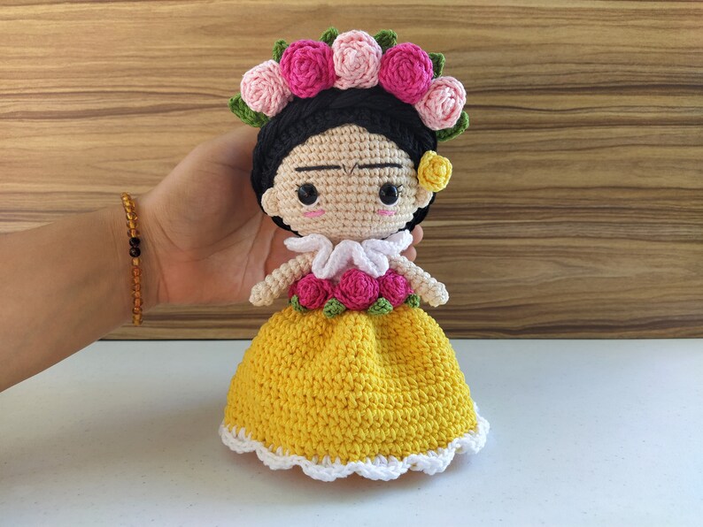 May include: A crocheted doll with black hair, a yellow dress, and a flower crown. The doll has a white collar and a pink flower on her ear.